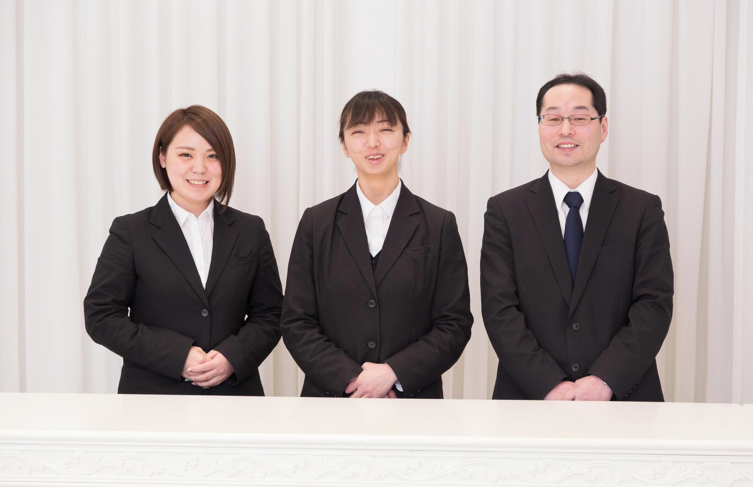 Three individuals in business attire standing in front of a white curtain backdrop.