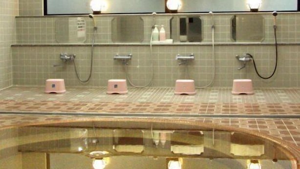 A public restroom with two sinks, each with a mirror and a light above. There are three pink soap dispensers and a hand dryer. The floor is tiled, and there is a glass partition in the foreground.