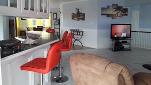 A modern living room with a beige sofa, a red chair, a kitchen area with white cabinets, and a television on a stand. - Rodney Heights Grande Splendour (Gros Islet)