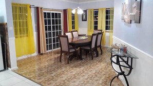 A dining area with a round table and chairs, a glass-top side table, and yellow curtains on the windows. - Rodney Heights Grande Splendour (Gros Islet)
