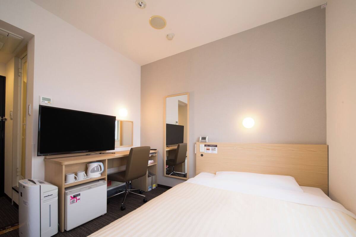 Habitación de hotel con cama, escritorio, televisor de pantalla plana, silla, kitchenette pequeño y espejo.