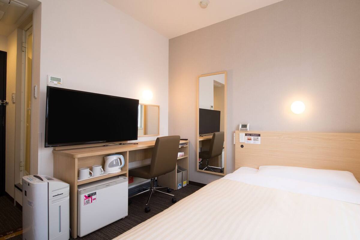 Habitación de hotel con cama, escritorio con televisor, silla, refrigerador pequeño, microondas, cafetera, espejo y termostato en la pared.