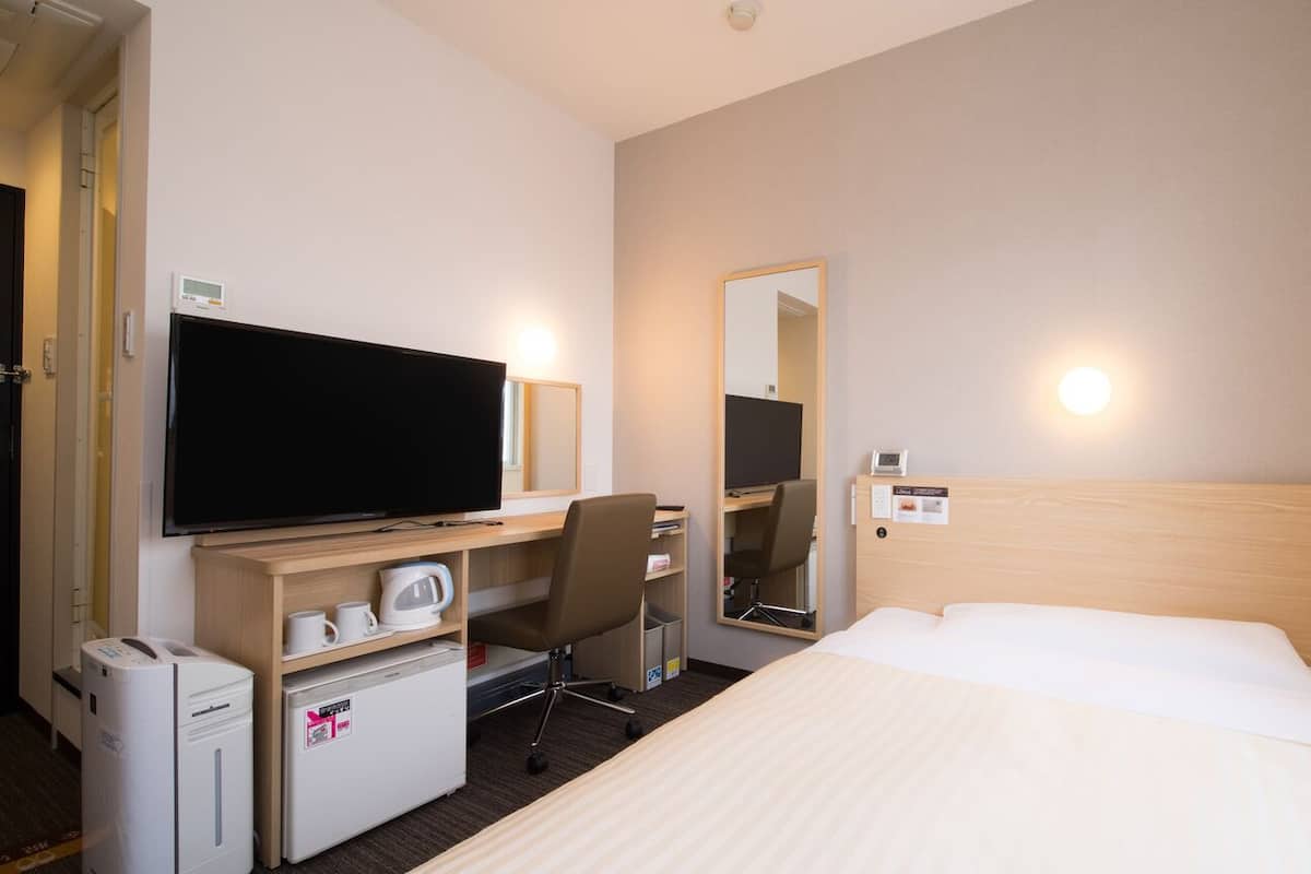Ein Hotelzimmer mit einem Bett, einem Schreibtisch mit Fernseher, einem Sessel, einem Minikühlschrank, einer Mikrowelle, einer Kaffeemaschine, einem Spiegel und einem wandmontierten Thermostat.