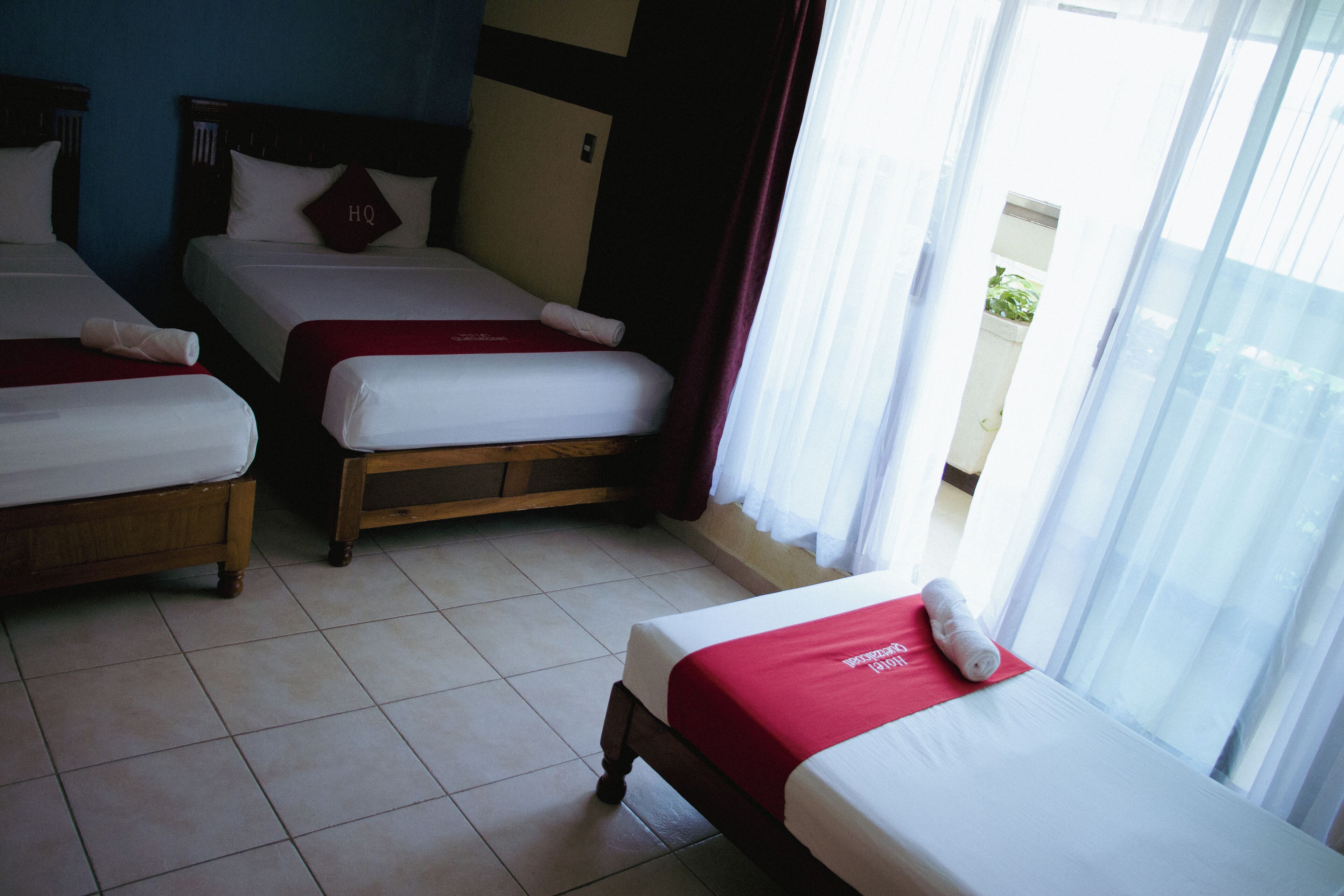 Habitación de hotel con tres camas, cada una con una manta roja y un cojín con la inscripción 'HQ'. El cuarto tiene piso de baldosas y una ventana con cortinas.