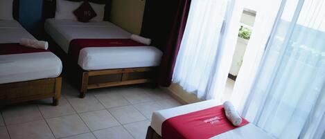 Habitación de hotel con tres camas, cada una con una manta roja y un cojín con la inscripción 'HQ'. El cuarto tiene piso de baldosas y una ventana con cortinas.