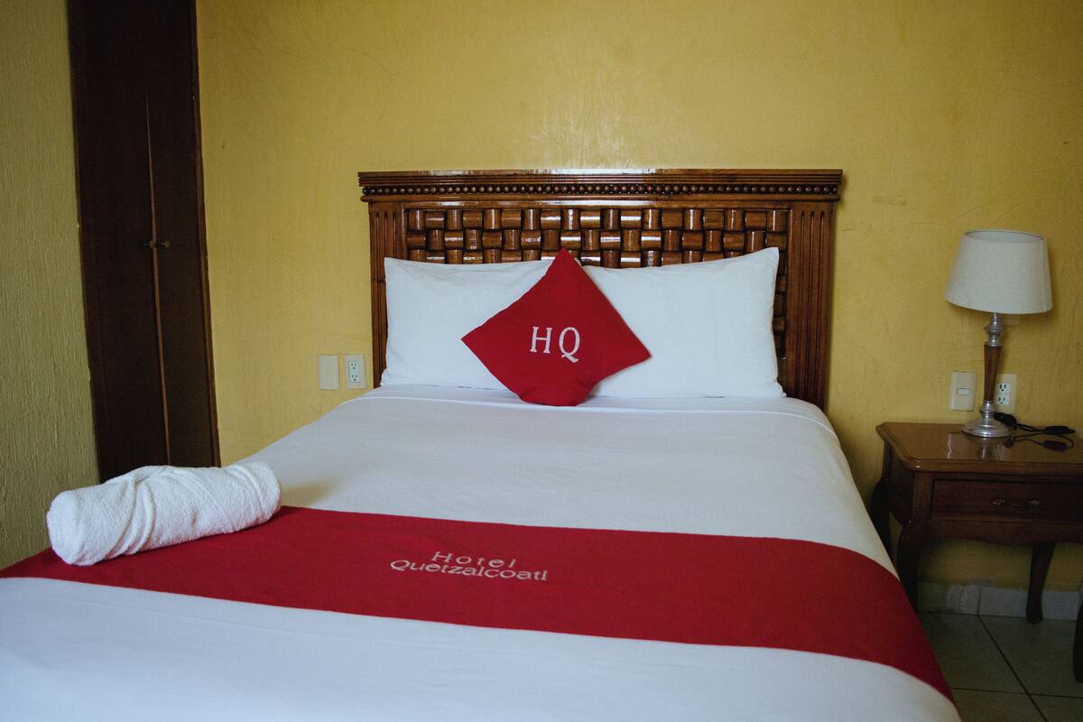 Une chambre d’hôtel avec un lit, une table de chevet avec une lampe, et une couvre-lit rouge et blanche ornée d’une broderie représentant le nom « Hôtel Queizalcoatl ».