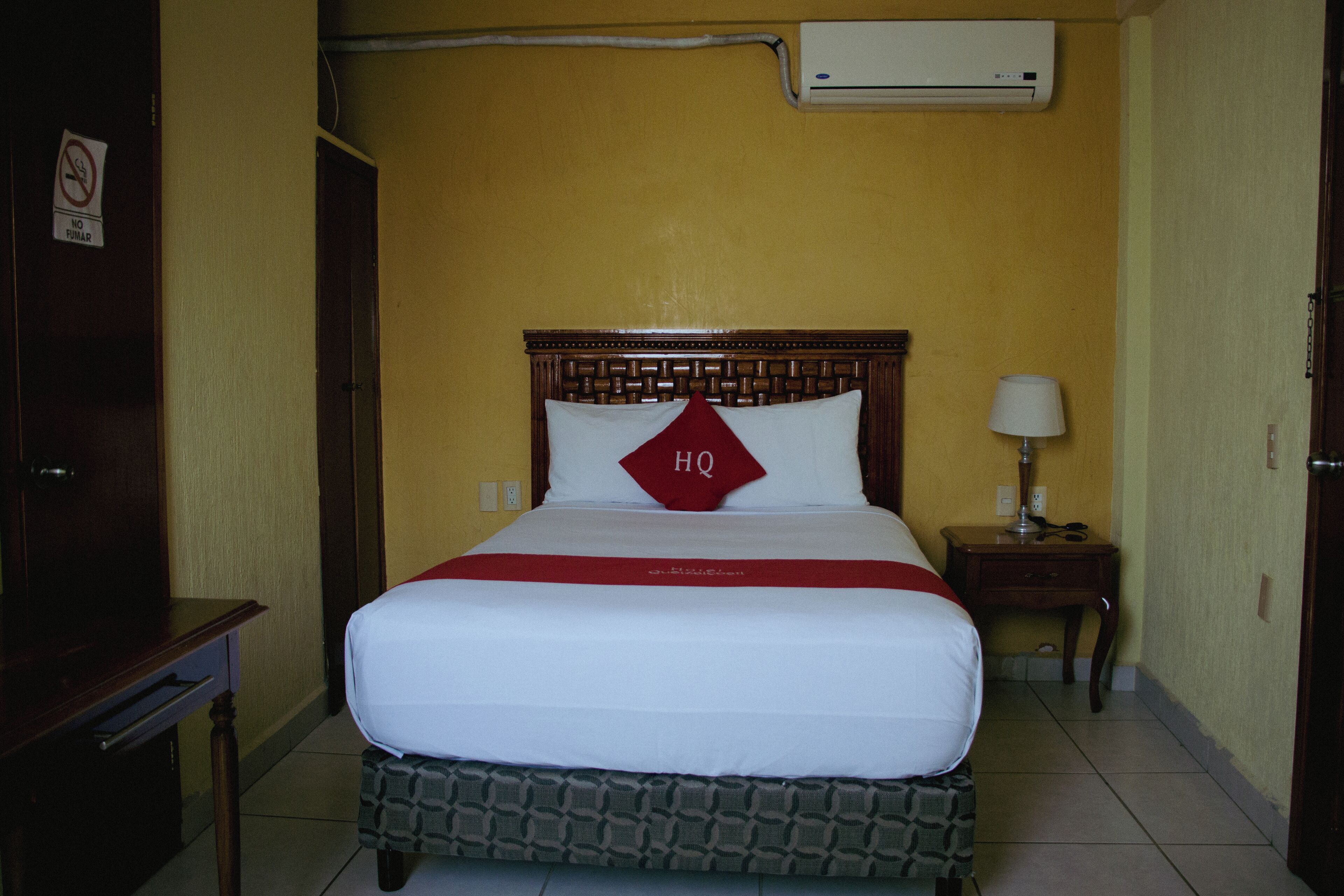 A hotel room with a bed, a nightstand with a lamp, and an air conditioning unit on the wall.