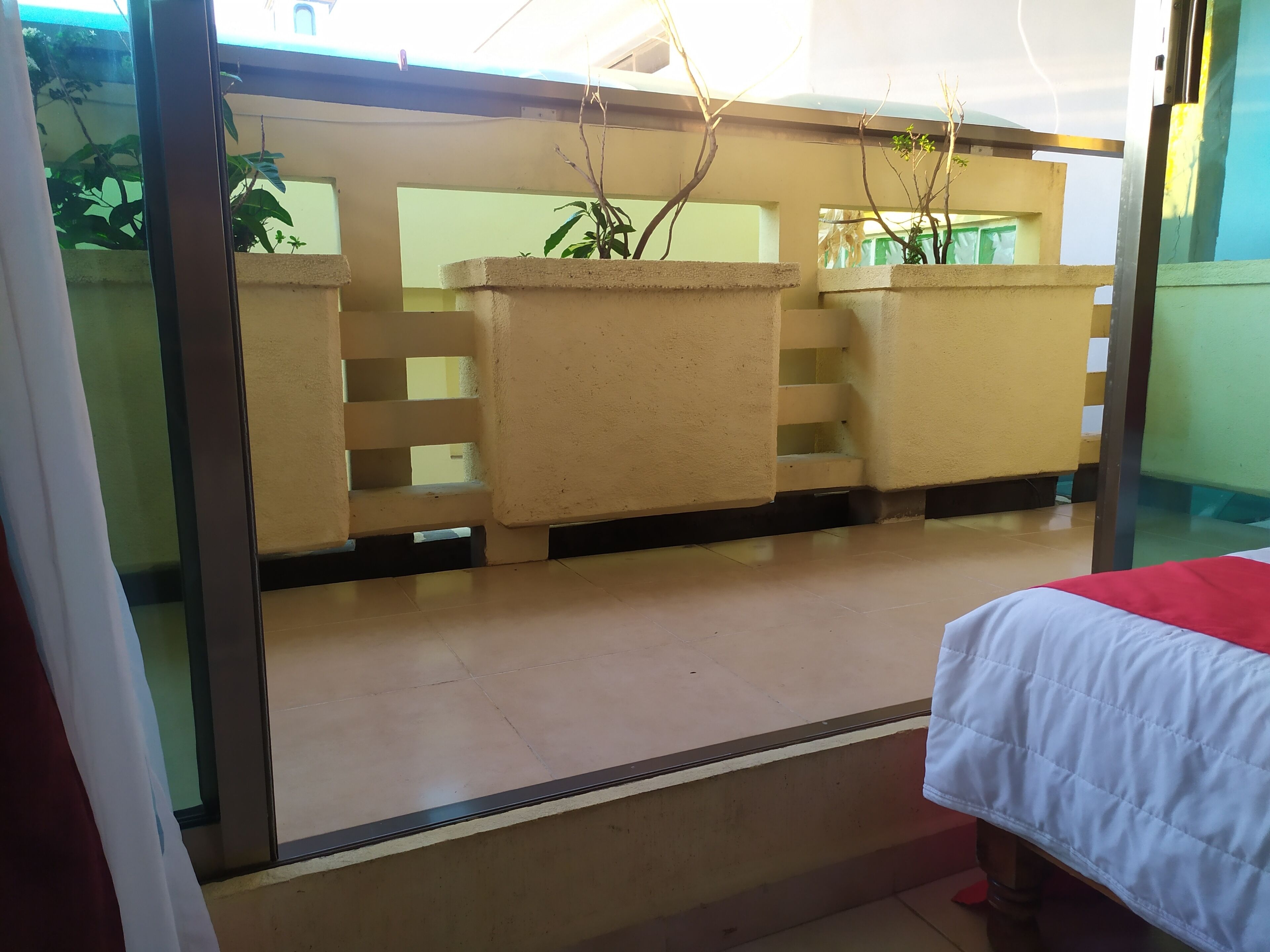 Quarto com cama, janela com vista para uma varanda e uma planta na varanda.