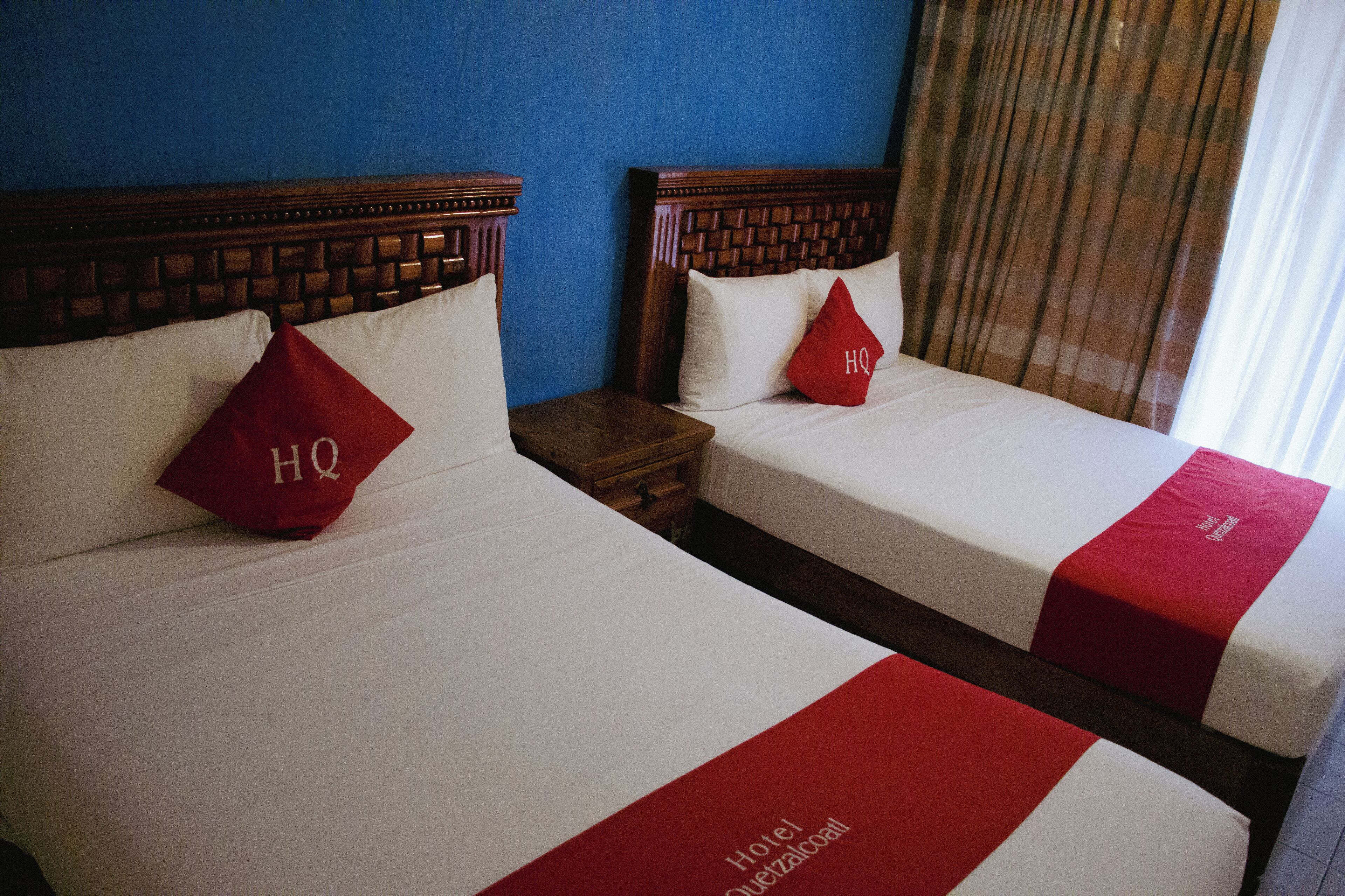 Dos camas de hotel con almohadones y colcha rojos, un cabecero de madera y una mesita de noche.