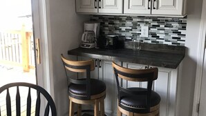 Une cuisine avec des meubles blancs, un dosseret carrelé et une machine à café. On y trouve deux chaises en bois, avec des assises noires et des pieds en métal.
