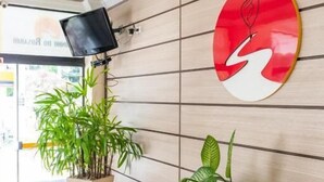 A reception area with a wooden wall, a red and white logo, a television, and potted plants. - Hotel Caminho do Rosário (Aparecida)