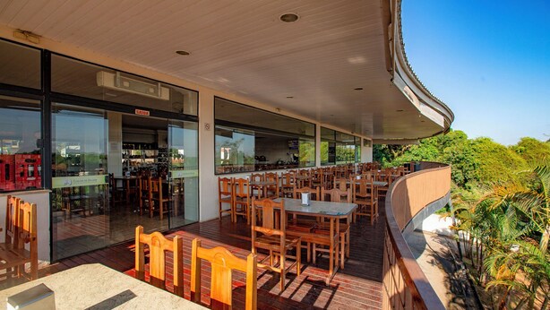 An outdoor dining area with wooden tables and chairs, a glass storefront, and a view of greenery.