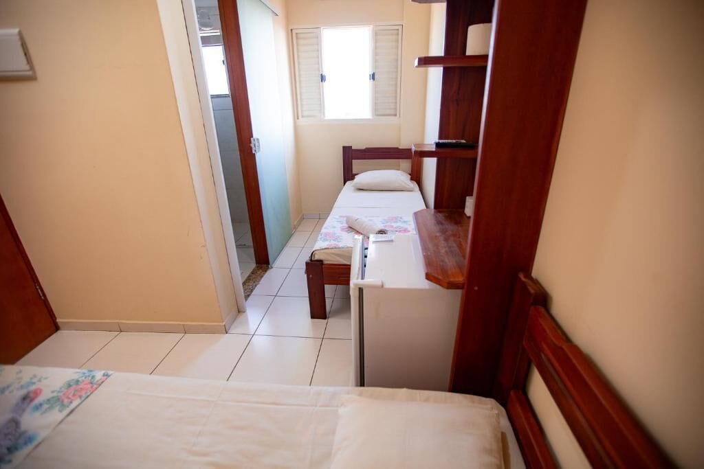 A narrow hallway with two beds, a wooden desk, and a window with shutters.