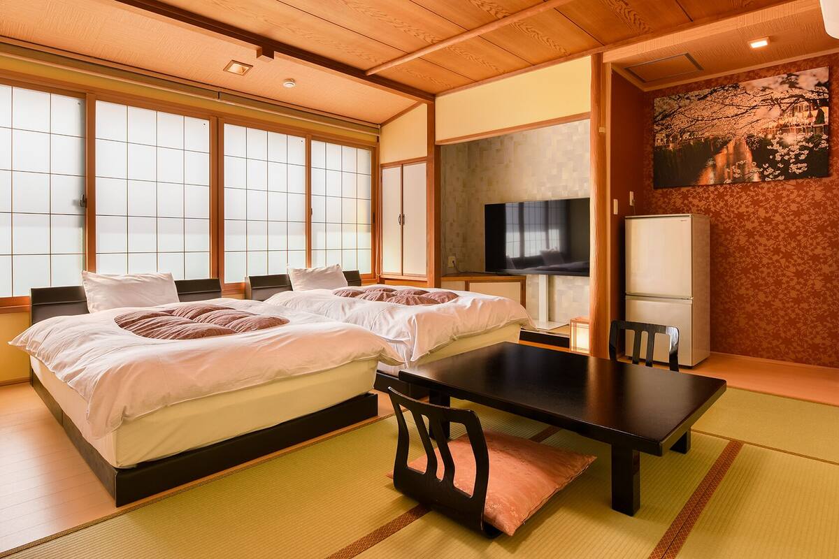 Ein traditioneller japanischer Raum mit Tatami-Bodenbelag, einem Bett mit weißer Bettwäsche, einem Fernseher und einem Kühlschrank.