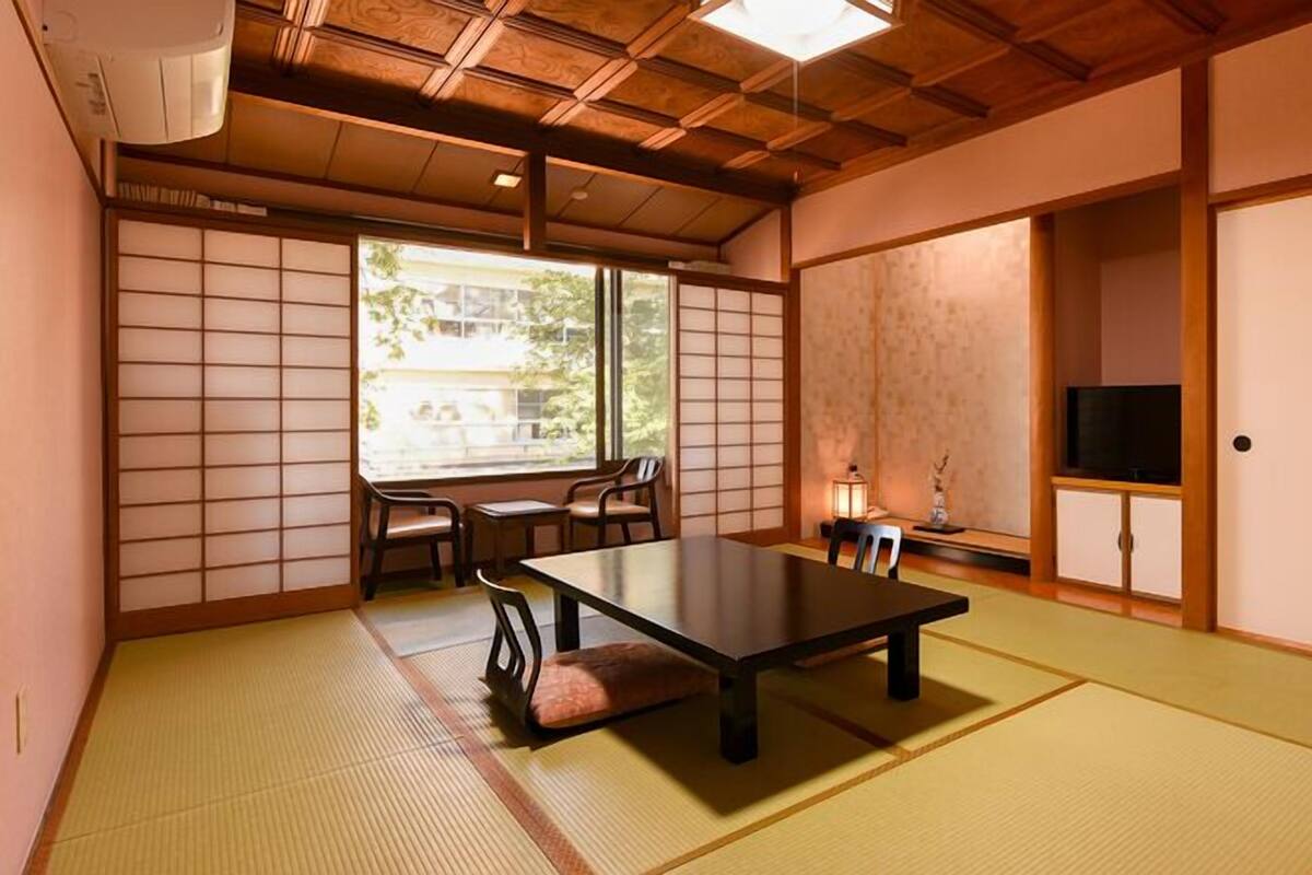 Ein traditioneller japanischer Raum mit Tatami-Bodenbelag, einem niedrigen Tisch, Stühlen, einem Fernseher und Schiebetüren.