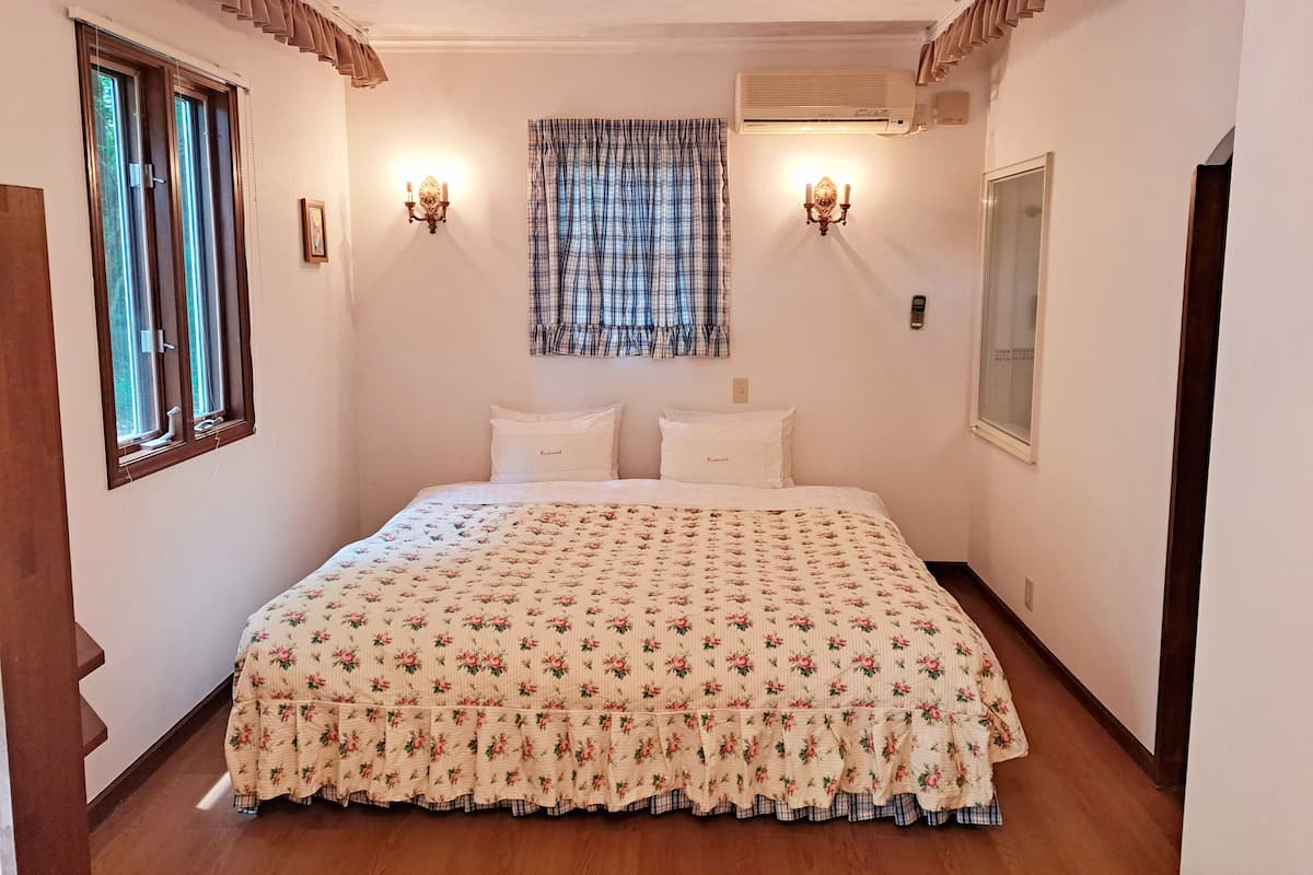 A bedroom with a floral bedspread, two bedside lamps, a window with curtains, and an air conditioning unit.