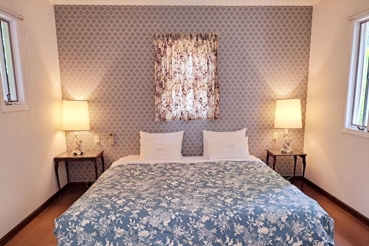 A bedroom with a patterned bedspread, two bedside lamps, a nightstand with a figurine, and a window with curtains.