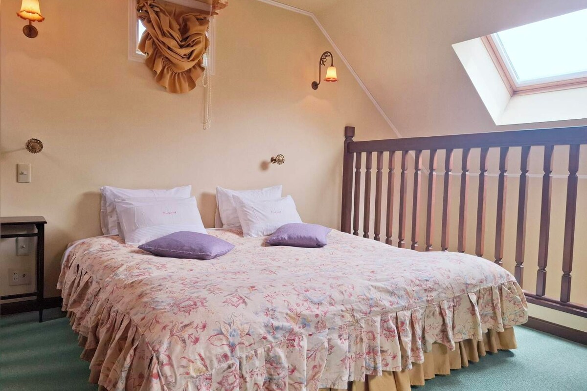 A bedroom with a sloped ceiling, a bed with floral bedding, two wall-mounted lamps, and a window with a view.