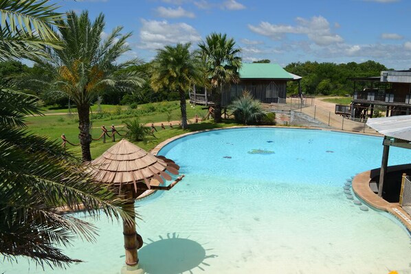 A swimming pool with a palm tree and a thatched-roof structure, surrounded by greenery and a building with a green roof. - Laguna Vista Tree House, Cloud Lane (New Braunfels)