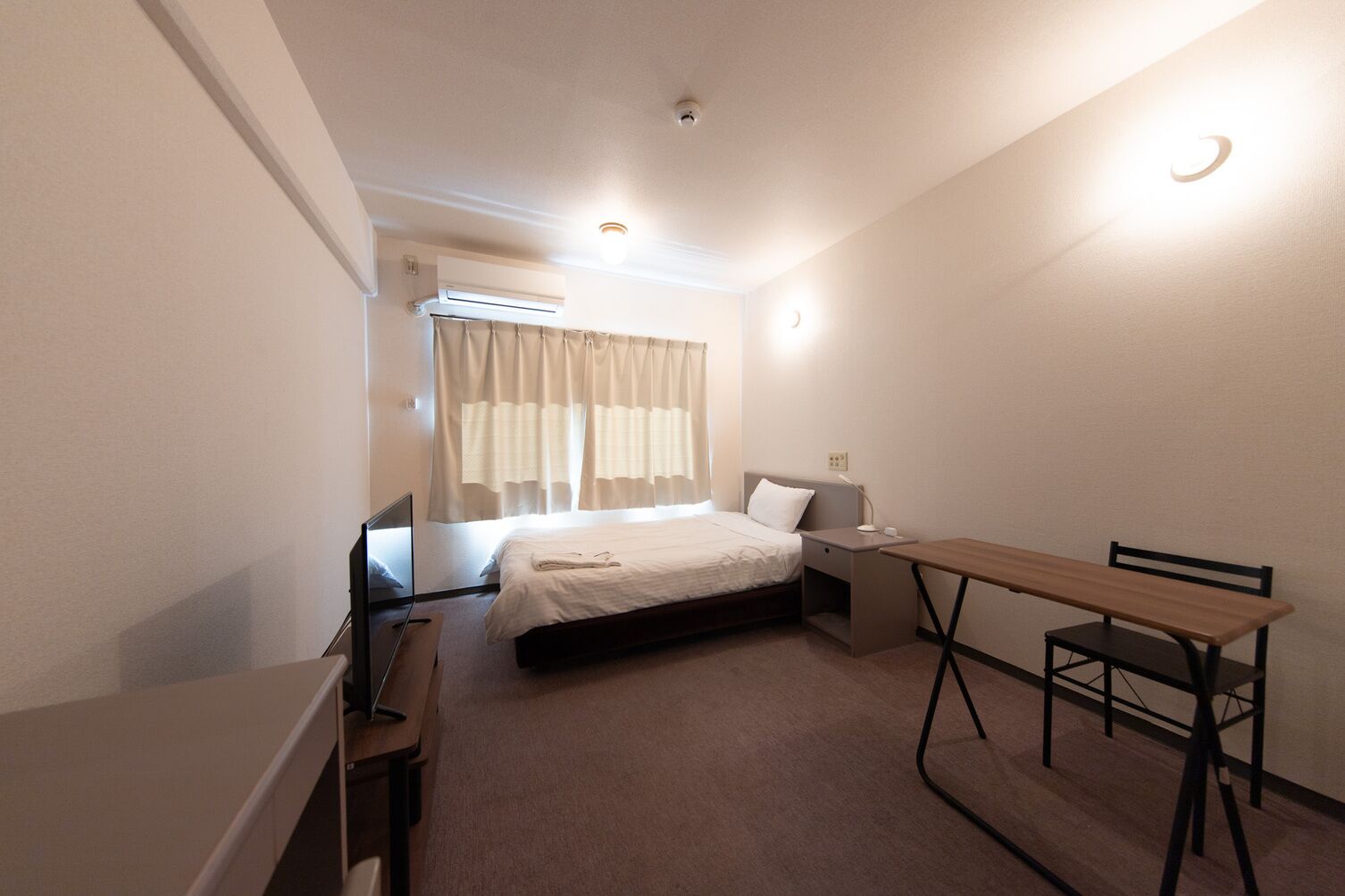 Een kleine, netjes ingerichte kamer met een bed, nachtkastje, bureau en stoel.
