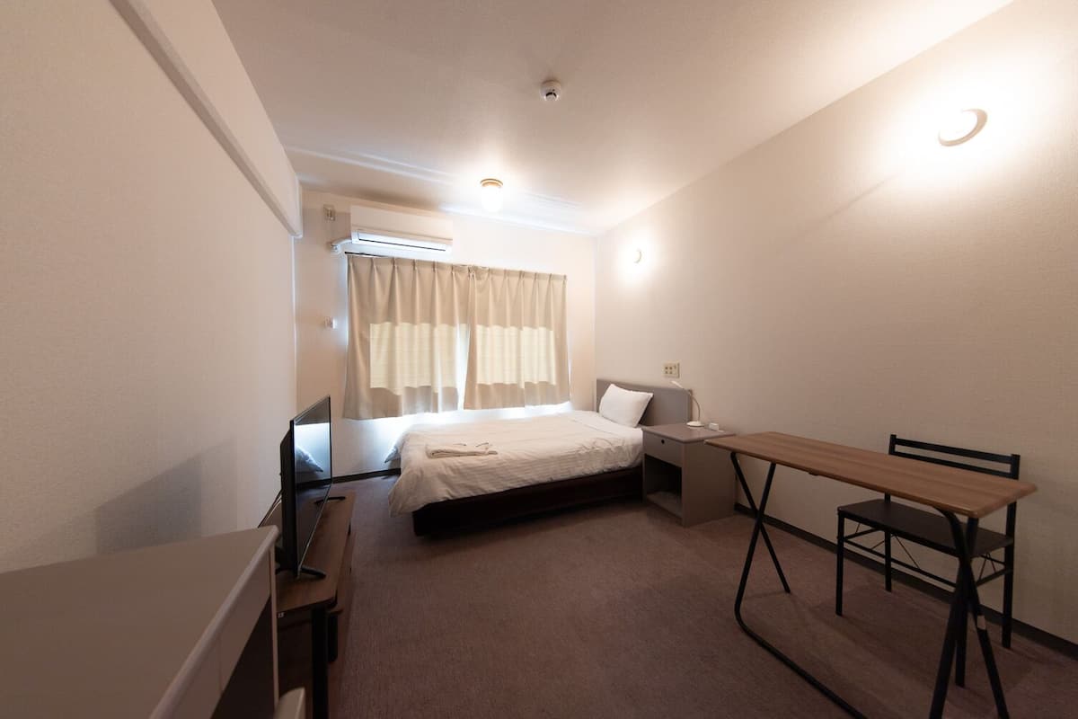 A small, neatly arranged room with a bed, bedside table, desk, and chair.