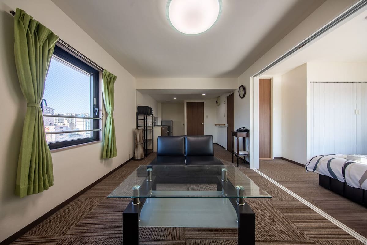 A hotel room with a glass-top table, a sofa, a bed, a view through a window, and a door leading to another room.