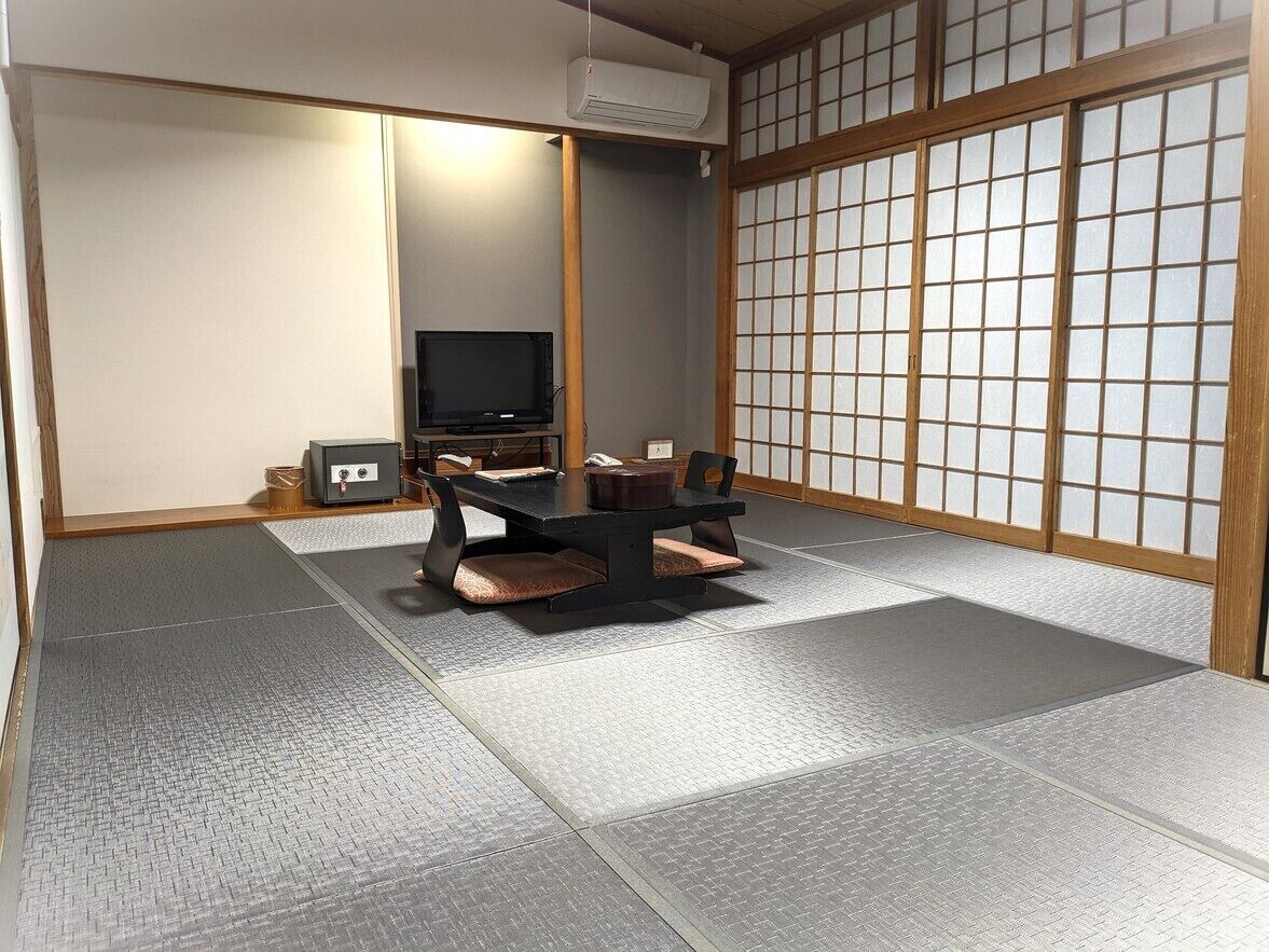 Ein Raum mit Tatami-Matten, einem niedrigen Tisch, Kissen, einem Fernseher und Schiebetüren.