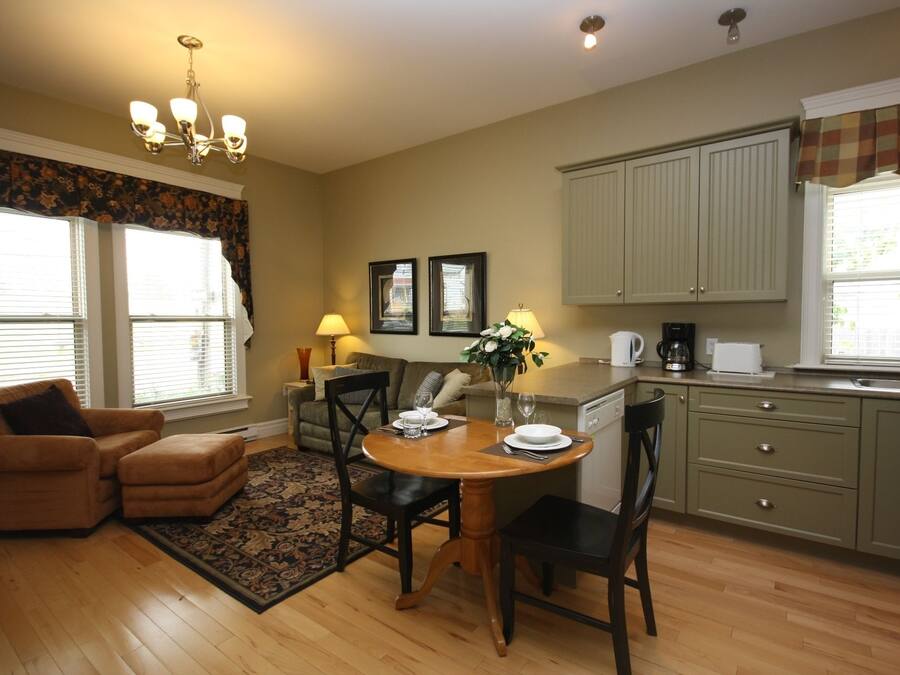 A cozy living room with a wooden dining table, a sofa, and a coffee table. There is a kitchen area with cabinets and a window.