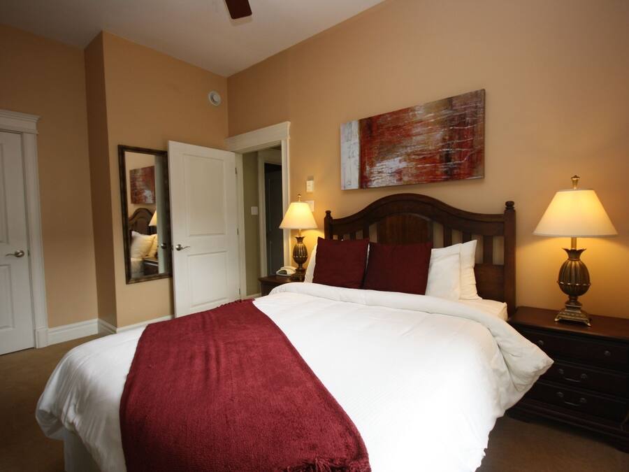 Quarto de hotel com uma cama grande, duas luminárias de cabeceira, um painel de cabeceira de madeira, um cobertor vermelho, um espelho e um quadro na parede.