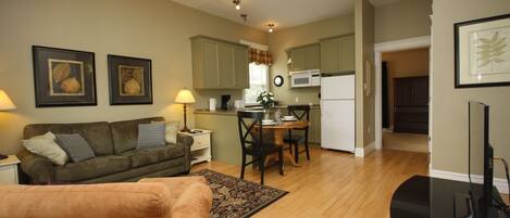 A living room with a sofa, a coffee table, and a TV. There is a kitchen area with cabinets and a microwave.