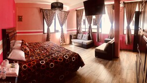 Habitación de hotel con una cama grande, un sofá y un televisor. Hay cortinas y una ventana con vistas.