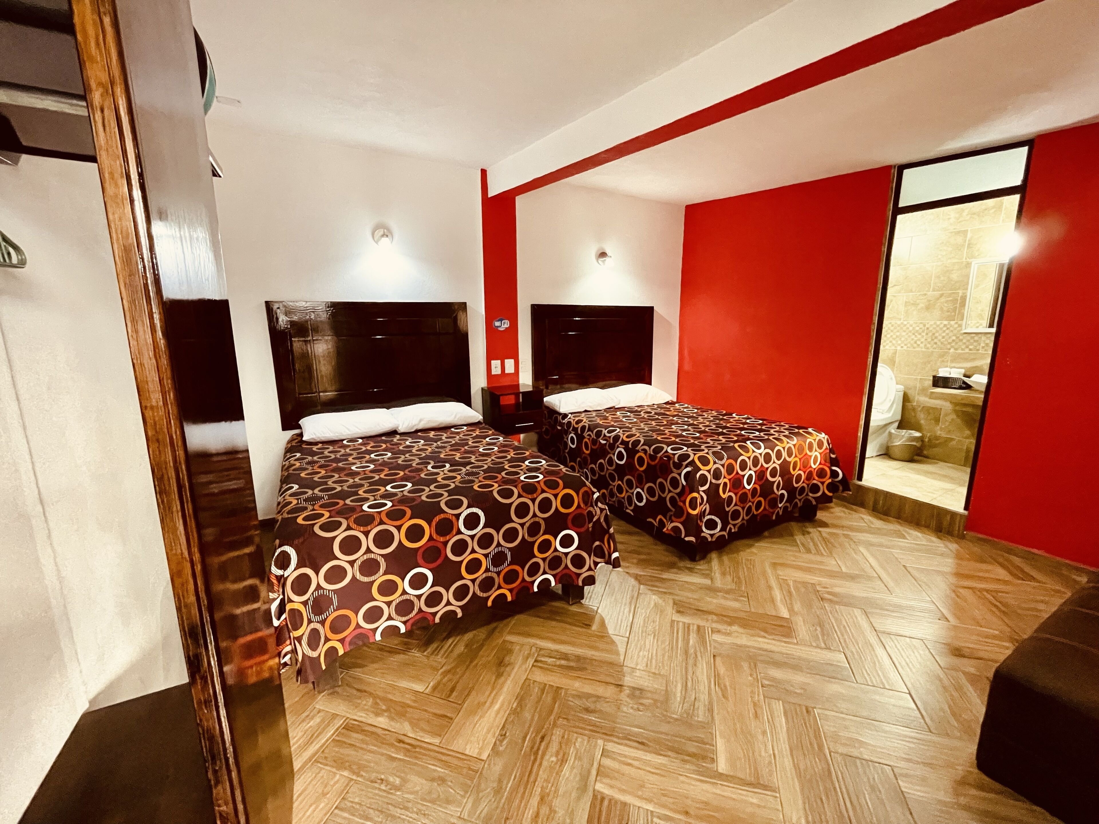 Habitación de hotel con dos camas, suelo de madera con diseño espiga, paredes rojas y un baño visible a través de una puerta de vidrio.