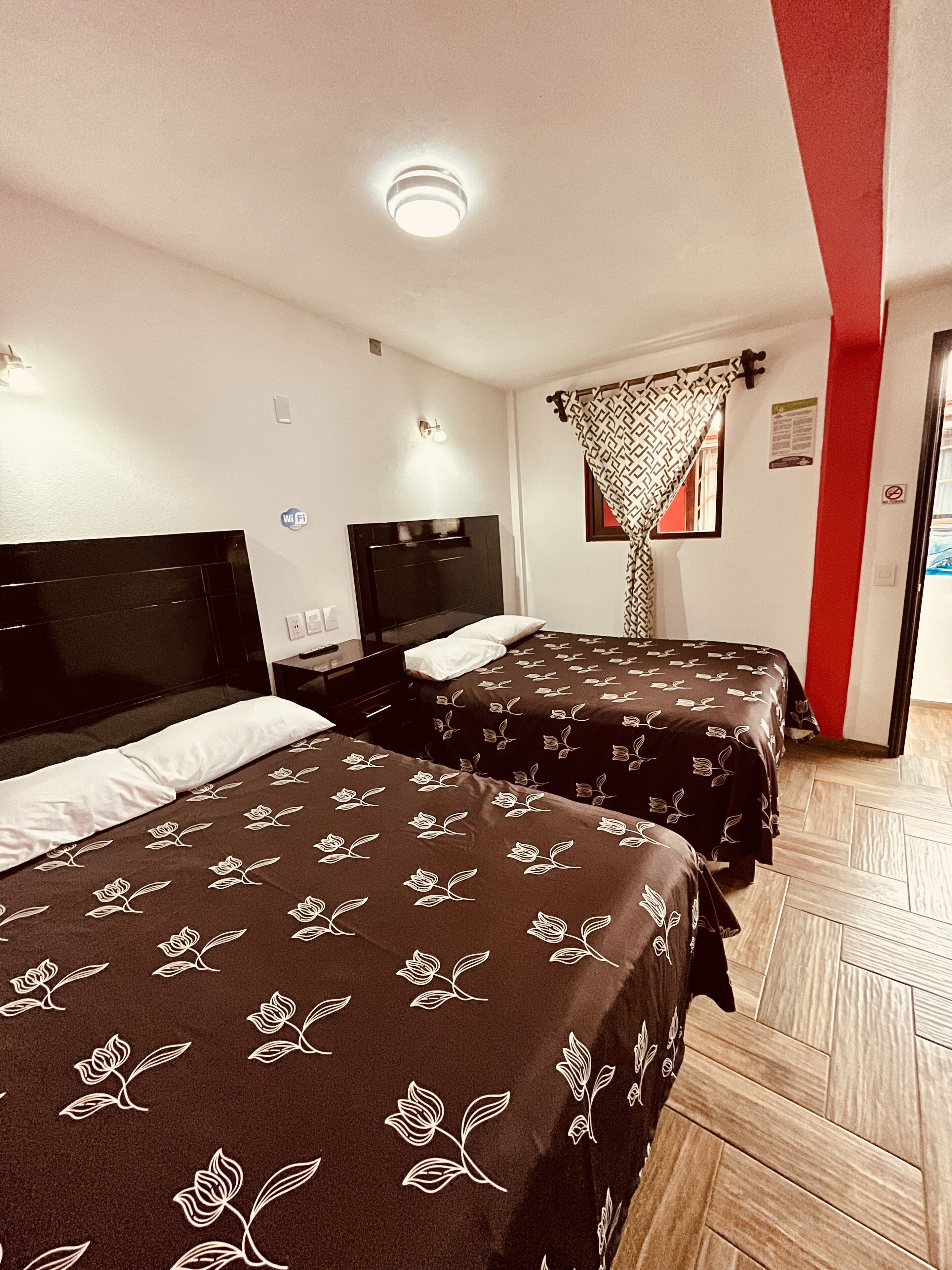 Habitación de hotel con dos camas, cada una con cabecero y colcha estampada. Se aprecia una lámpara en el techo, una luz de pared y una ventana con cortina decorada.