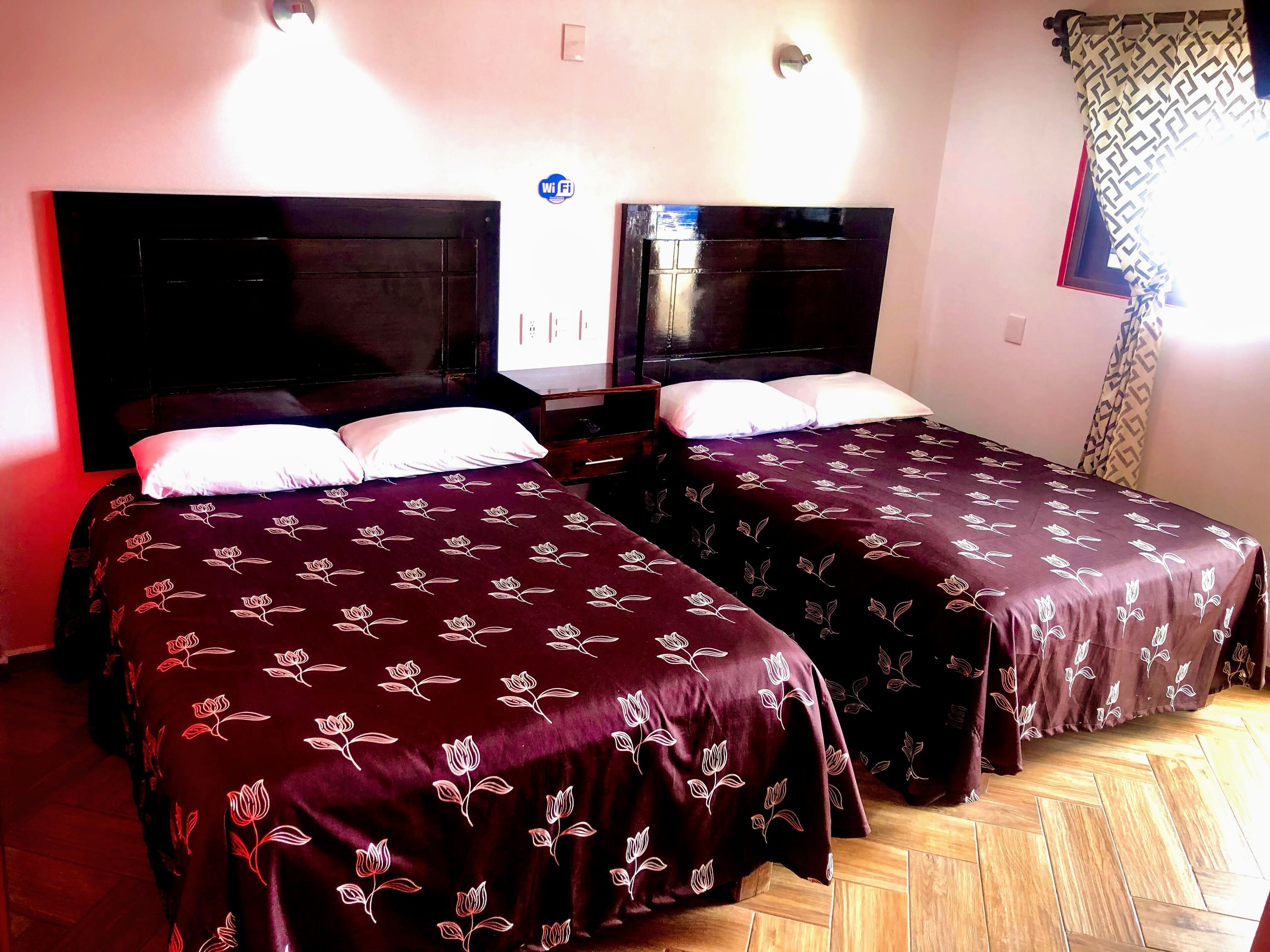 Habitación de hotel con dos camas, cada una con una colcha floral oscura y almohadas blancas. El suelo es de madera y hay un aplique de pared sobre las camas.