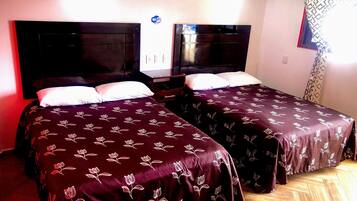 Habitación de hotel con dos camas, cada una con una colcha floral oscura y almohadas blancas. El suelo es de madera y hay un aplique de pared sobre las camas.
