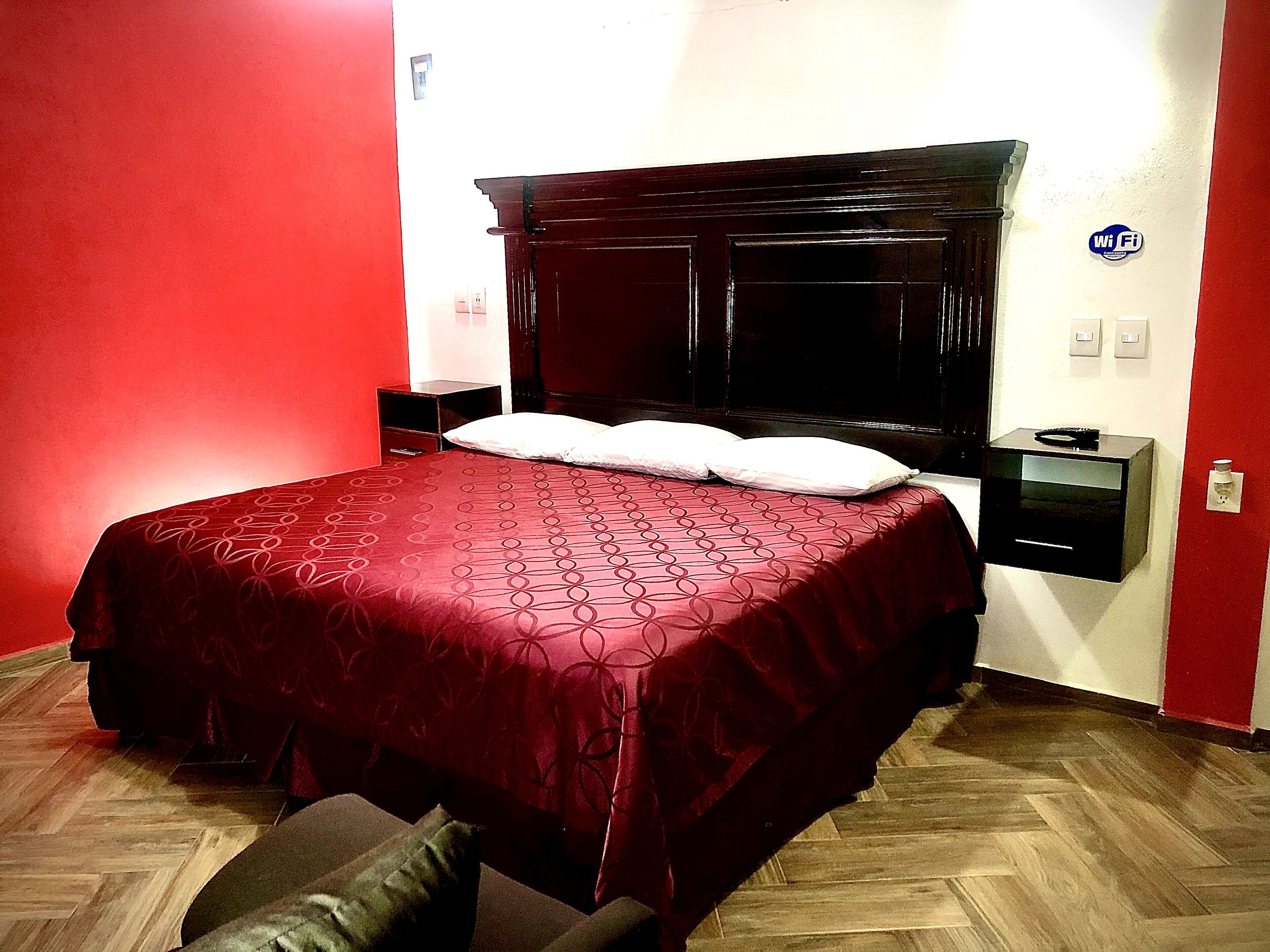 A bedroom with a large bed, a nightstand, a red wall, and a Wi-Fi sign.