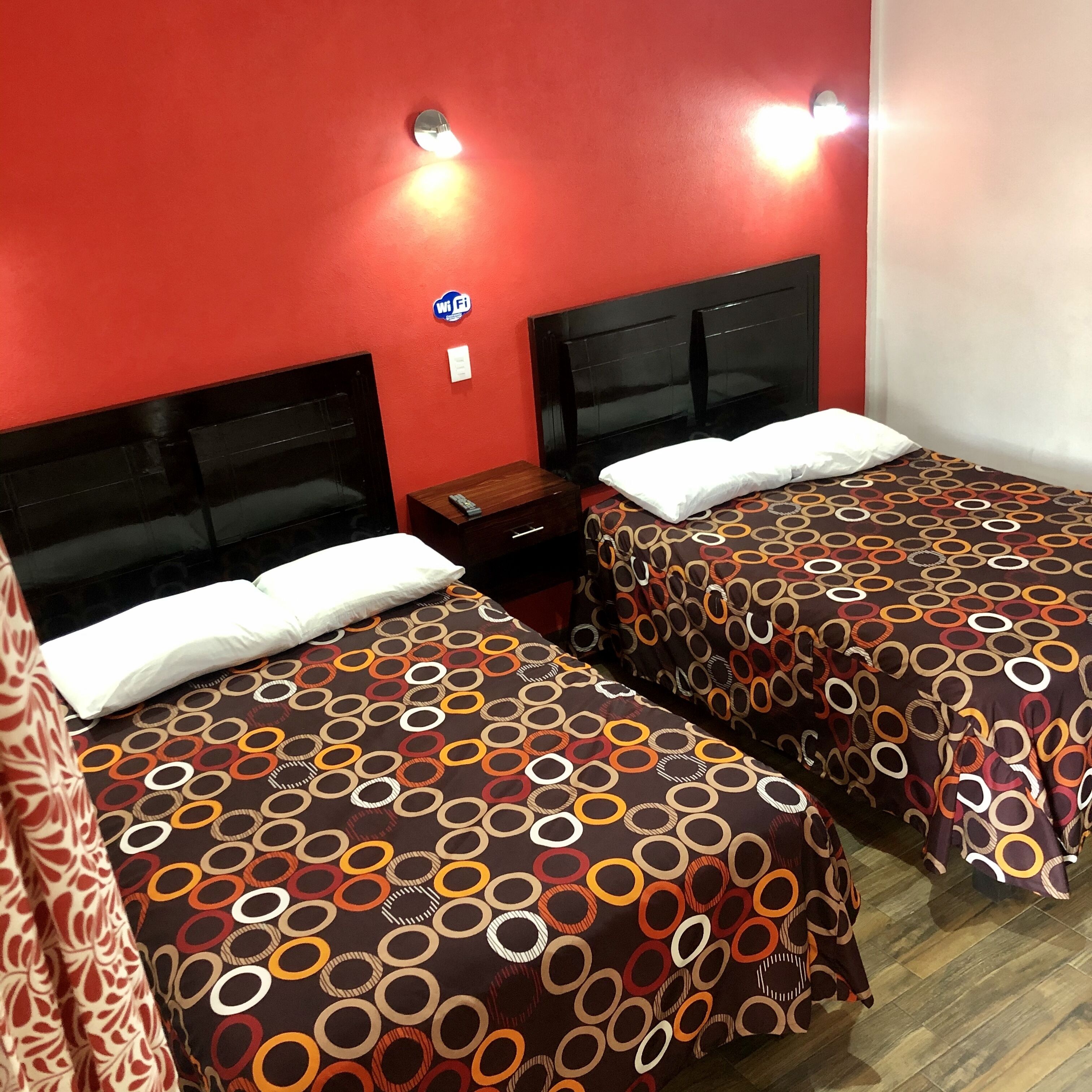 A hotel room with two beds, each with a patterned bedspread, a wooden nightstand, a TV, and a Wi-Fi sign on the wall.