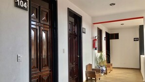 Un pasillo con puertas de madera, indicaciones con números, un extintor y un banco.