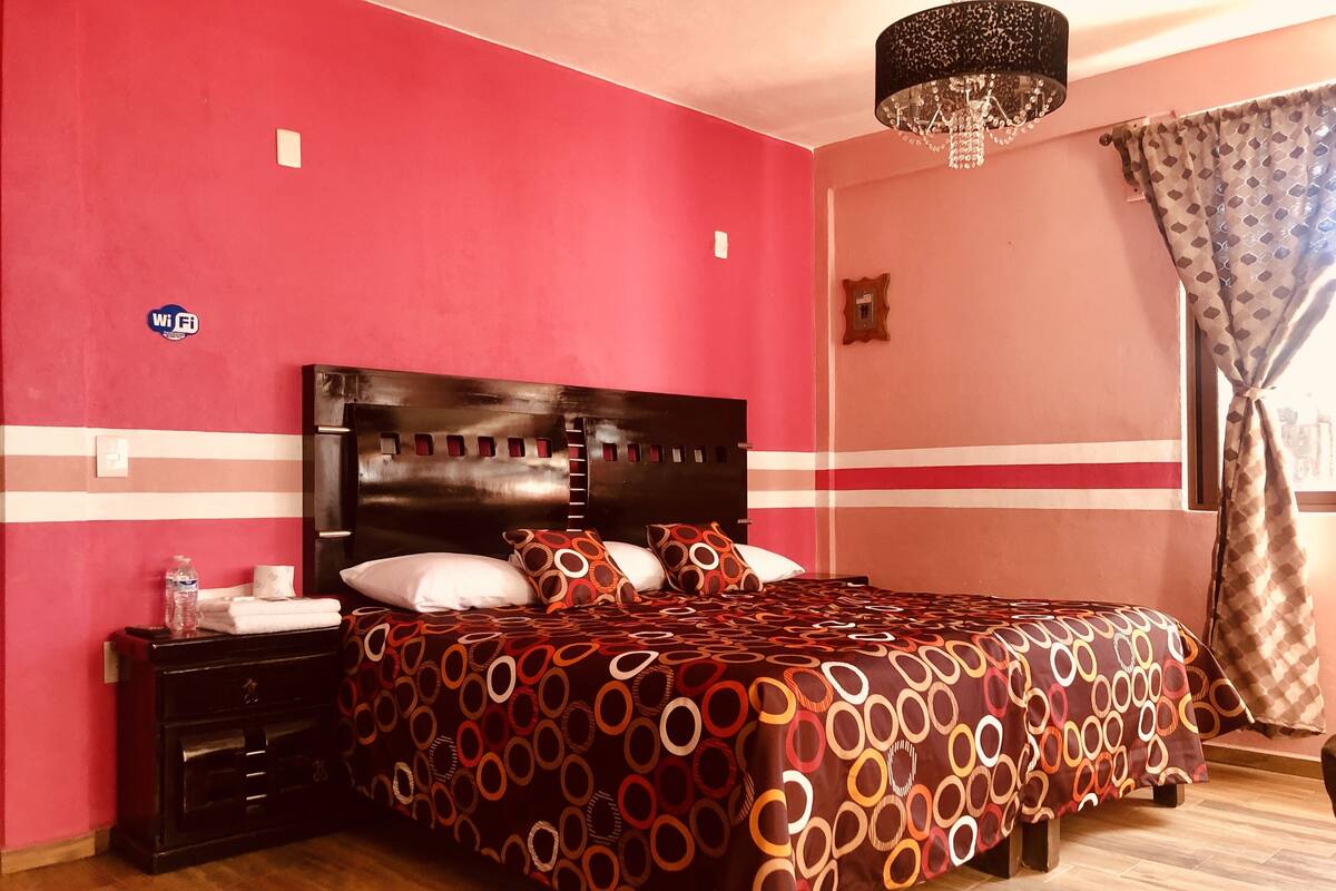A bedroom with a red wall, a wooden headboard, a patterned bedspread, a nightstand with a bottle, and a chandelier.