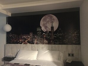 A bedroom with a cityscape mural on the wall, a bed with white bedding, a bedside table, and a hanging light fixture. - Hotel La Luna (Mexico City)