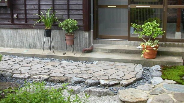 A stone pathway leading to a building with a wooden facade and a glass door. There are three potted plants on the steps. - Minshuku Jinya (Fukui)