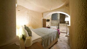 A bedroom with a stone wall, a bed with a patterned bedspread, a nightstand, a lamp, a wooden bench, and a stone fireplace. - Arte Cave Hotel (Ürgüp)