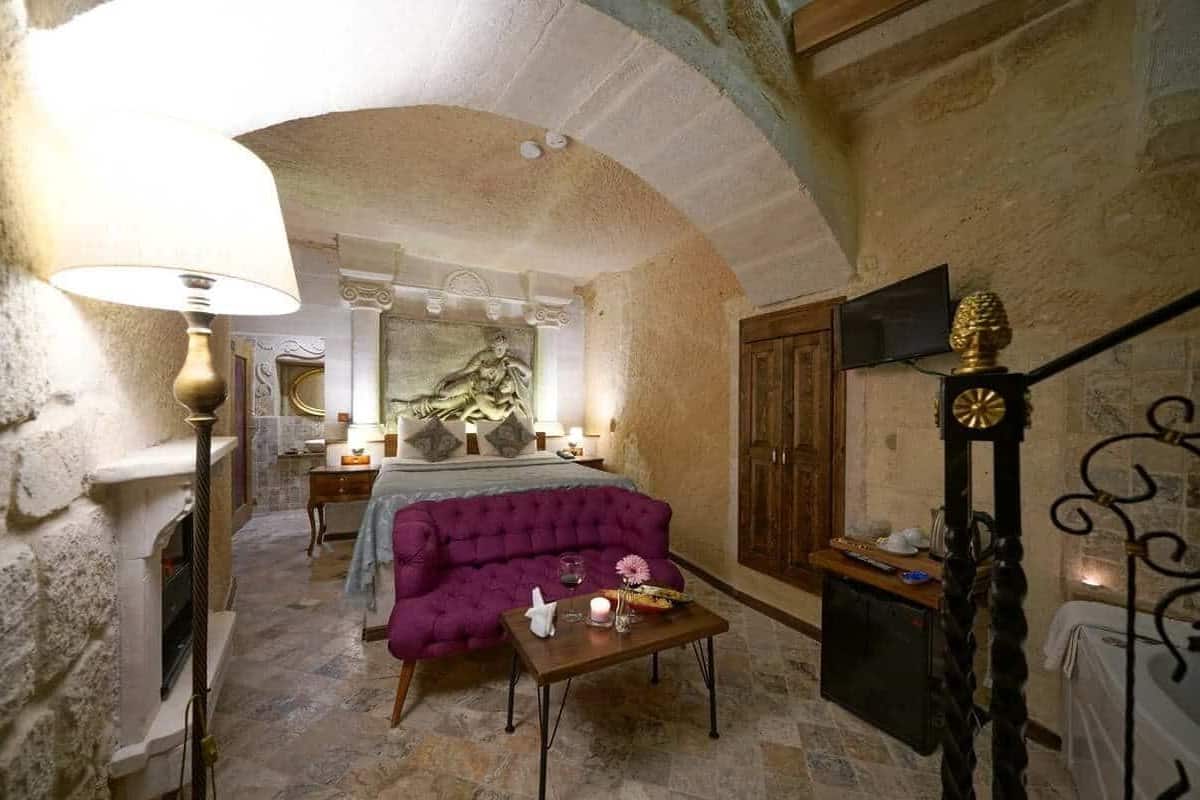 A room with a fireplace, a purple sofa, a wooden table with candles, and a bed with a headboard.