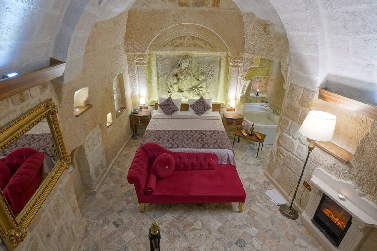 A bedroom with a red sofa, a bed with two pillows, a nightstand, a lamp, and a fireplace.