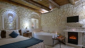 A bedroom with a stone wall, a fireplace, a large bed, a bathtub, and a television. - Arte Cave Hotel (Ürgüp)
