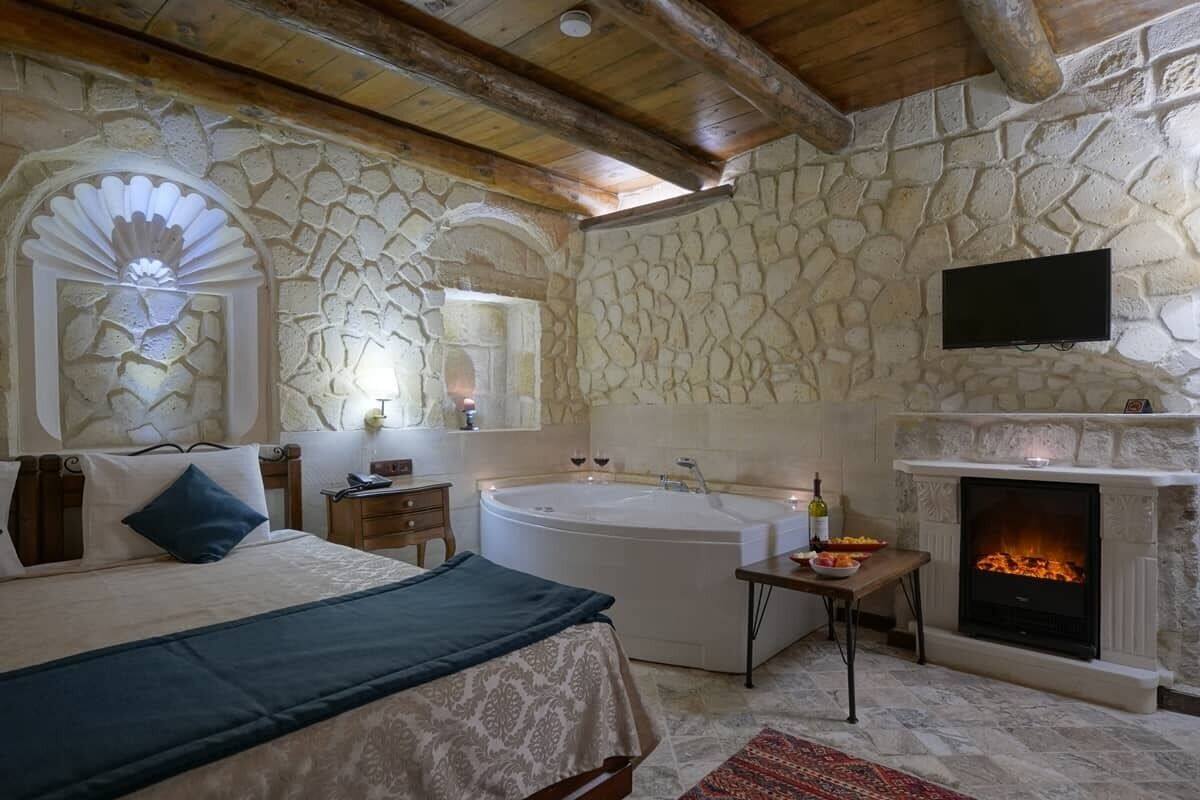 A bedroom with a stone wall, a fireplace, a large bed, a bathtub, and a television.