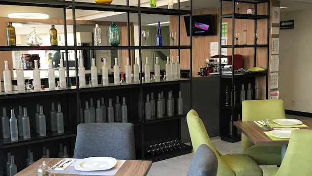 Un espace repas moderne avec une table en bois dressée pour deux personnes, entourée de chaises rembourrées de couleur grise et verte. Une cloison en métal noir, dotée d’étagères, expose diverses bouteilles en verre et des objets décoratifs.