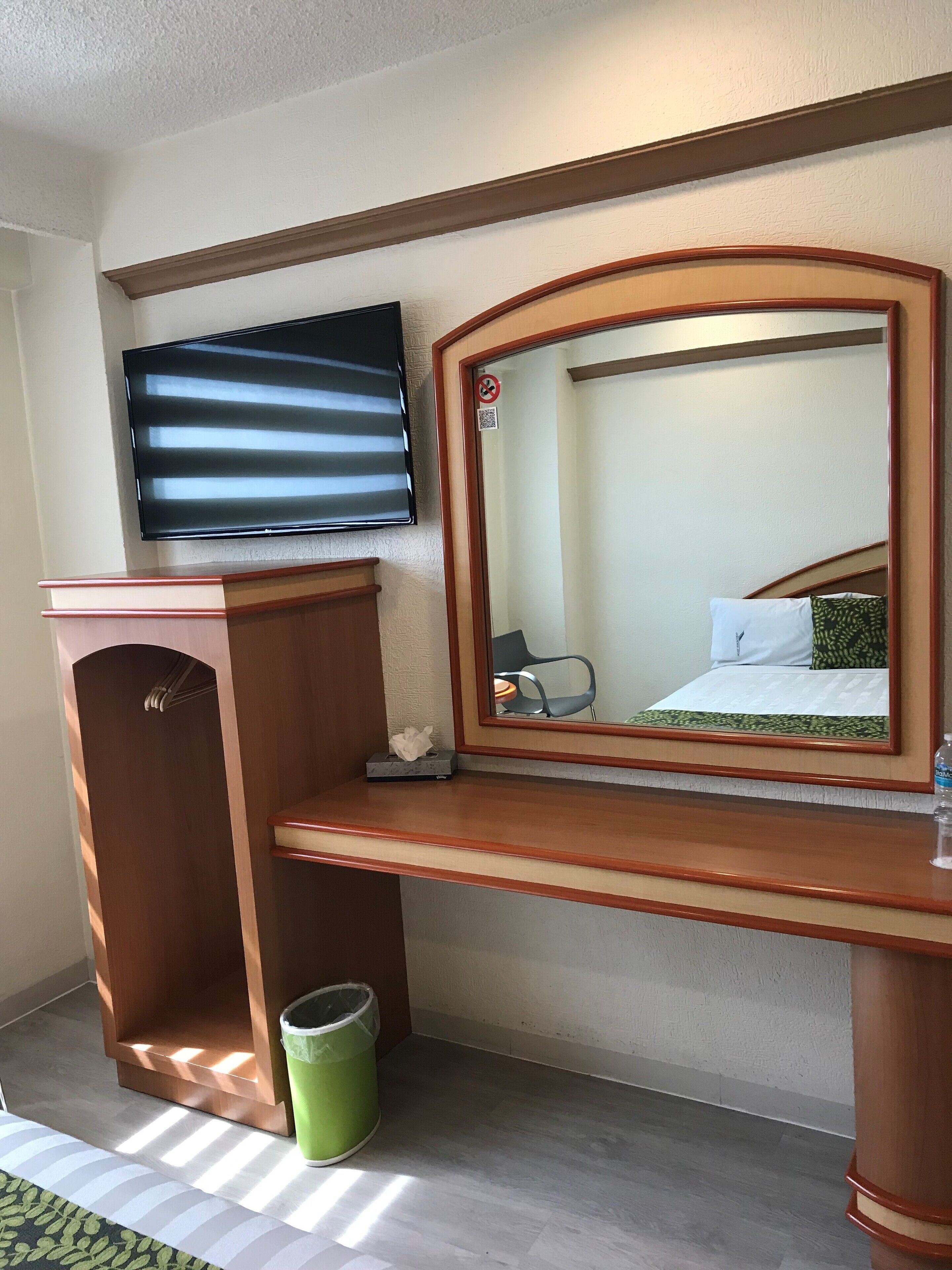 Ein Hotelzimmer mit einem Holztisch, einem an der Wand montierten Fernseher, einem Spiegel, einem Mülleimer und einem Bett mit einer grün-weißen Bettdecke.