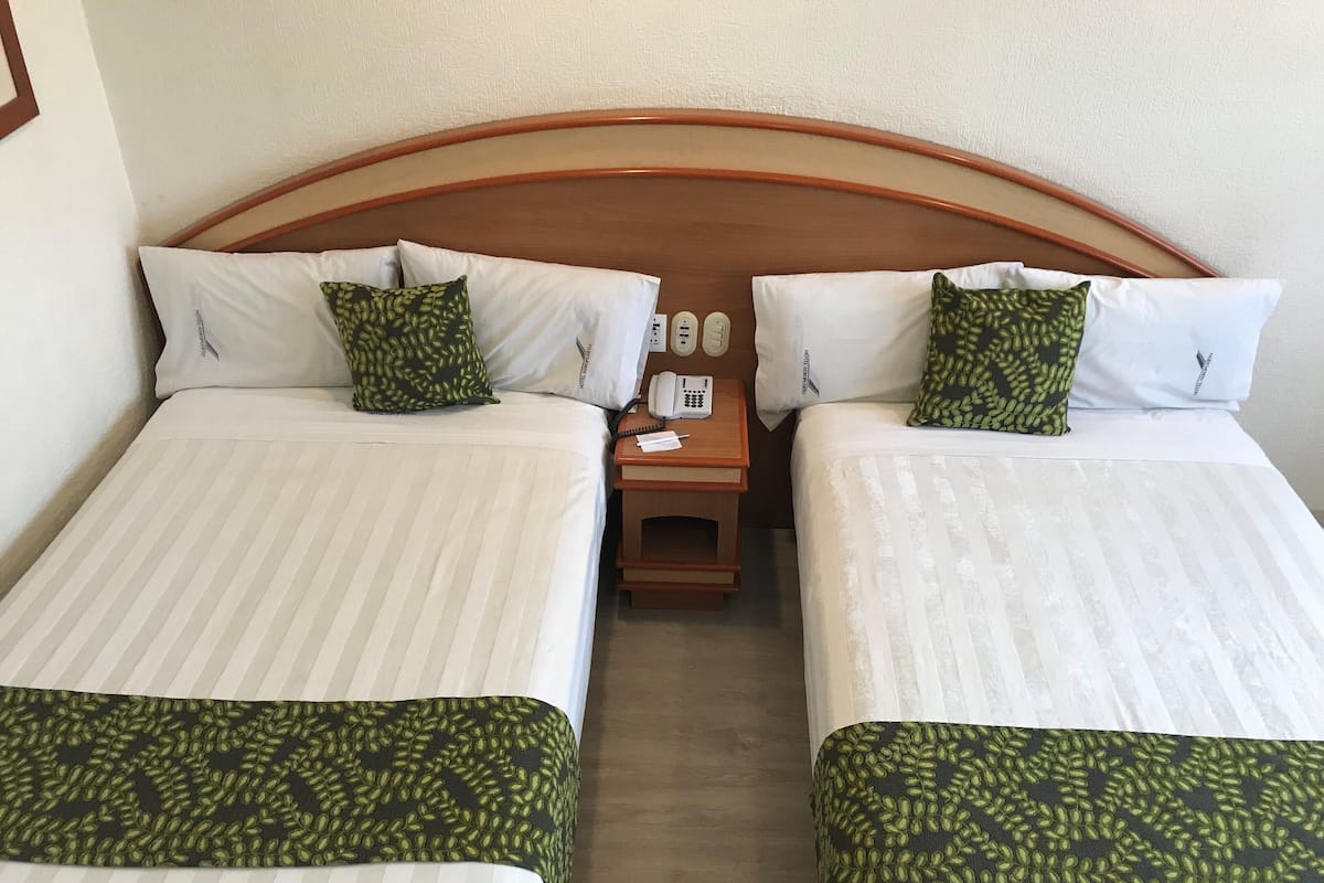 Habitación de hotel con dos camas, cada una con ropa de cama blanca y almohadas con estampado verde. Tiene un cabecero de madera, una mesita de noche con un teléfono y una lámpara, y una pequeña mesa con un papel y un bolígrafo.