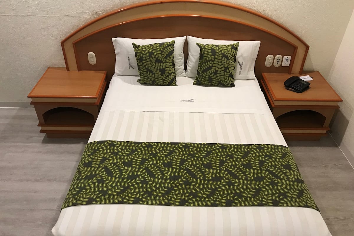 Habitación de hotel con cabecera de madera, dos almohadas blancas, colcha de cama con estampado verde y manta a rayas. Se observan dos mesitas de noche de madera, una con un teléfono y una lámpara.